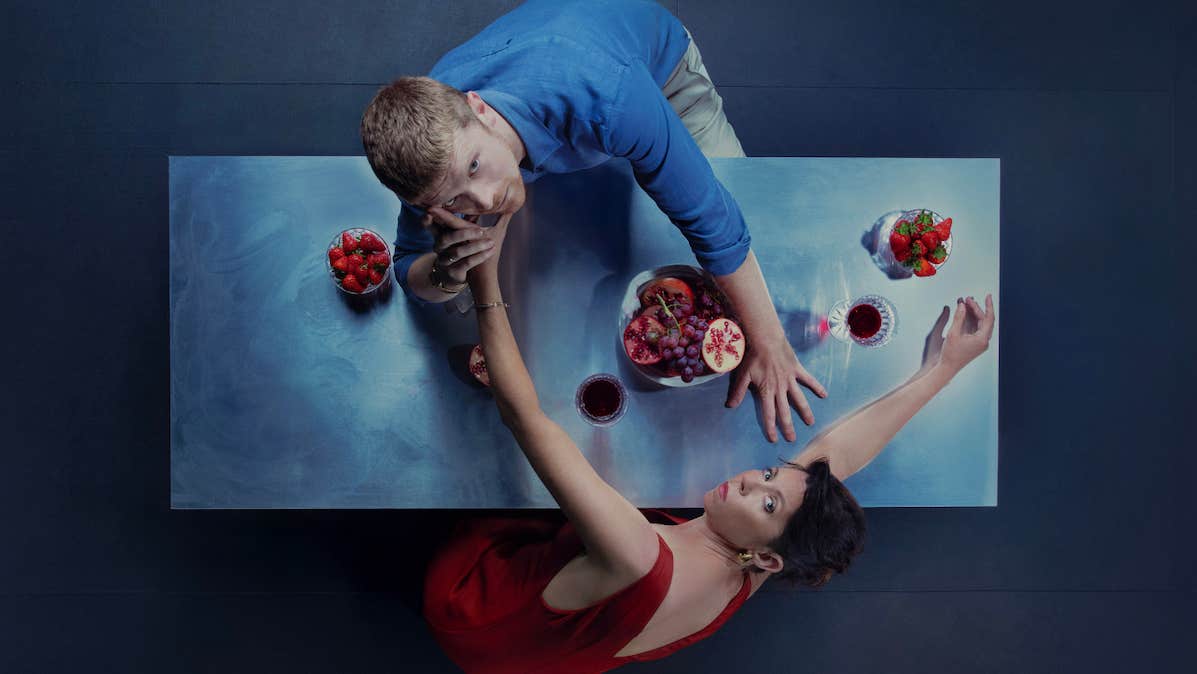 Viewed from above, a man and woman are reaching across a dinner table to each other, looking up at the viewer.