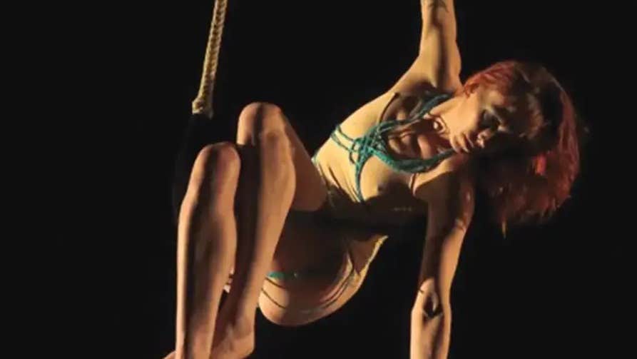 A person is hanging from a rope by one hand, against black background.
