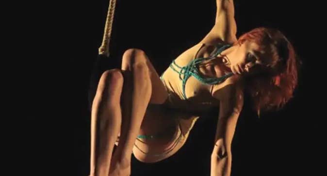 A person is hanging from a rope by one hand, against black background.