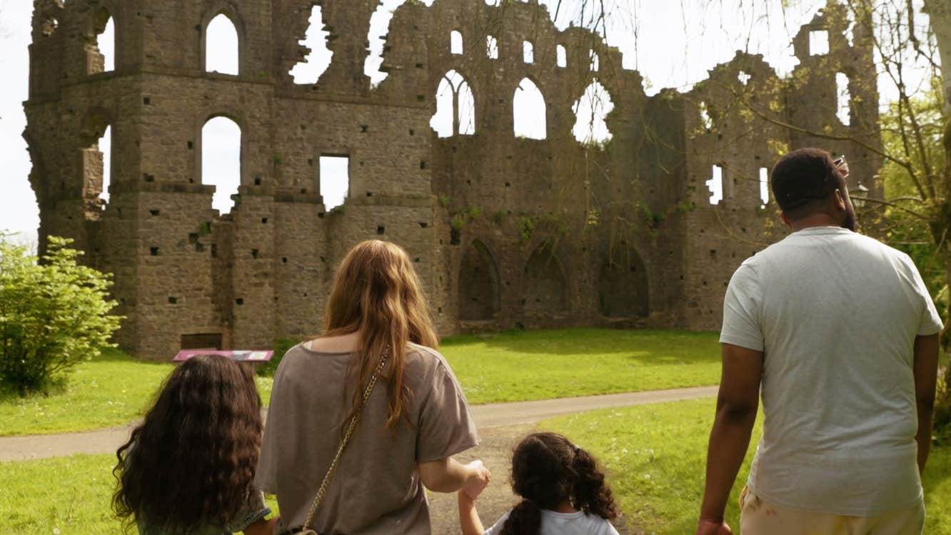 Four people holding hands walking through a garden towards an old stone ruin