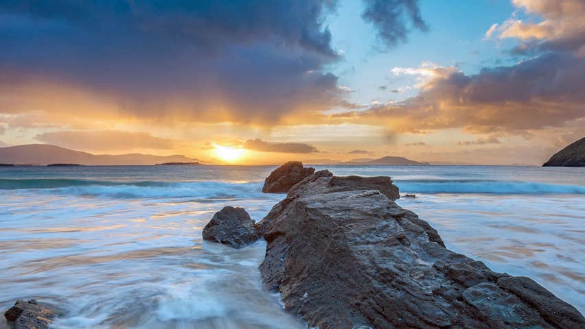 An atmospheric photo of a beach scene at sunrise or sunset with foaming sea around a large rock on a sandy beach