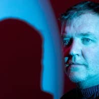 A serious looking man lit in blue with shade of his head cast to one side of him.