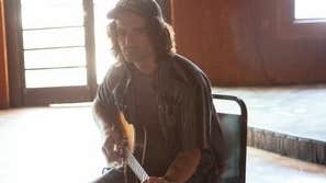 A serious looking man seated on a chair with a guitar with back to the light from a doorway