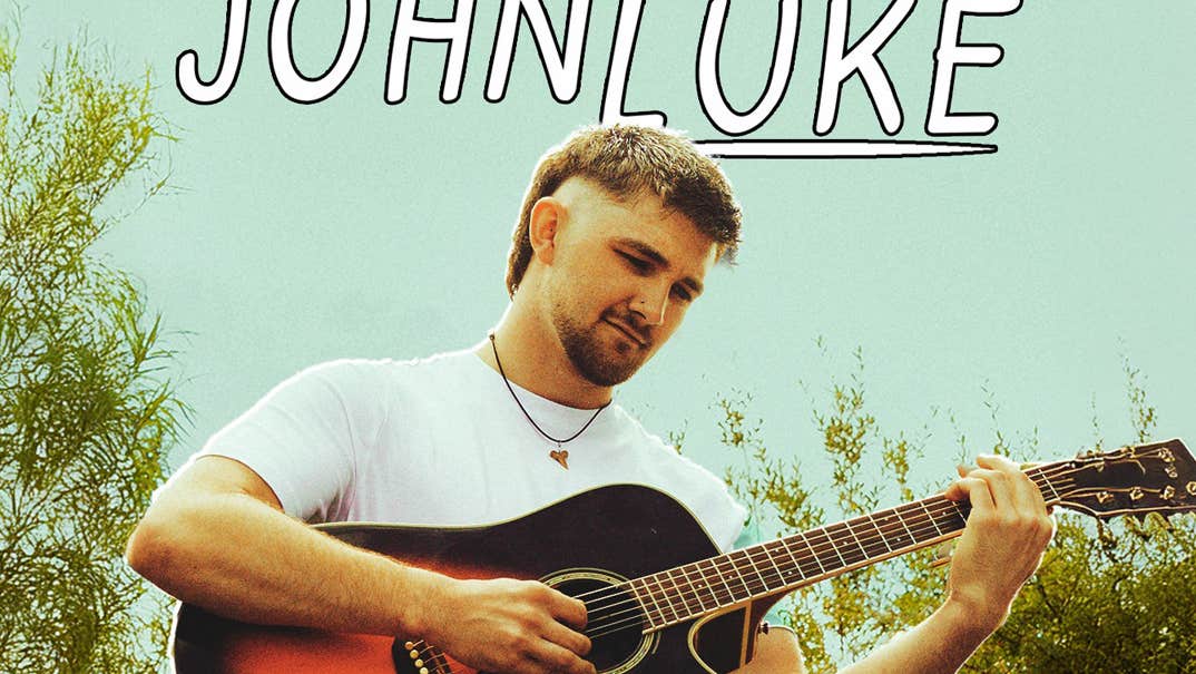 Singer/songwriter and musician Johnluke, a slightly smiling man is seated on a rock playing a guitar, surrounded by greenery