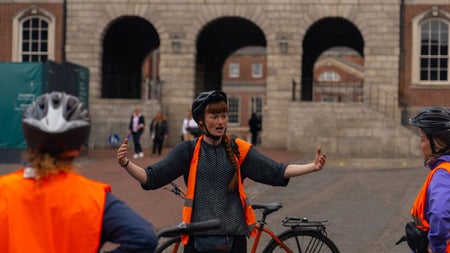 A lady standing in front of a bicycle wearing an orange high visibility vest and helmet