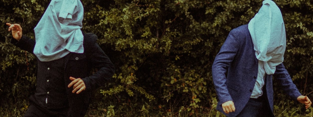 2 people outdoors with hedge in background, standing apart with large, pale blue garments over their heads.