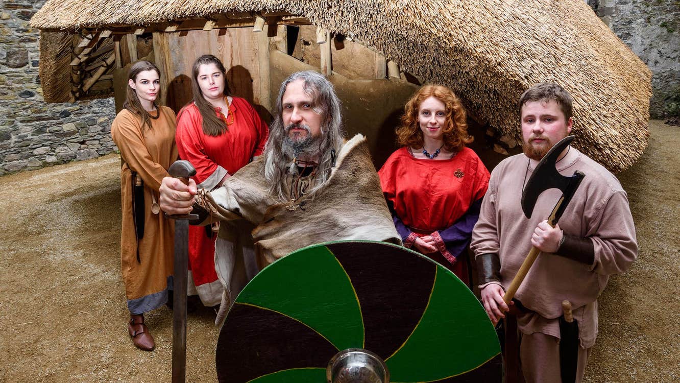 People dressed at Vikings holding weapons and a green and black shield outside a thatched replica Viking house