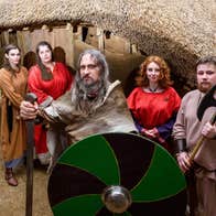 People dressed at Vikings holding weapons and a green and black shield outside a thatched replica Viking house