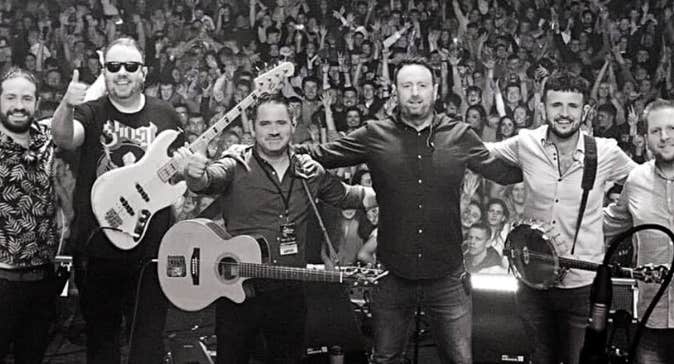 A line of 6 smiling men on a stage with their backs to a large audience.