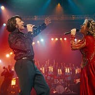 A man and woman singing with passion into mics, facing each other on a stage.