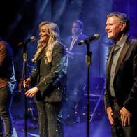 3 people in a line on a stage each singing into a mic on a stand with musicians behind.