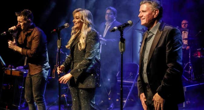 3 people in a line on a stage each singing into a mic on a stand with musicians behind.