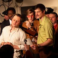 A man is pointing a shotgun at a bound man while holding a sheet of paper. Someone is holding a microphone towards them and there are other men in the background.