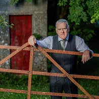 A rather cross looking older man in shirt, tie and waist coat is leaning his arms on a rusty gate