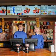 Two people standing behind a wooden counter with packets of sweets a popcorn machine and light up sign advertising cinema snacks