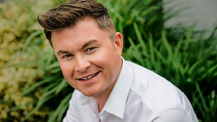 A slightly smiling man in white shirt with grasses in the background.