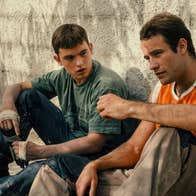 A boy in his late teens and a young man are sitting against a wall.