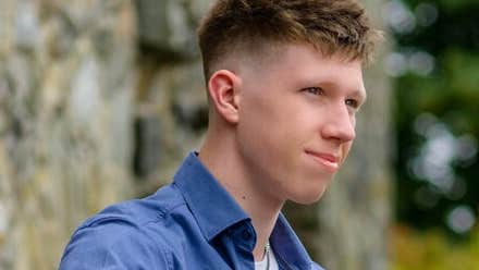 A gently smiling young man with blurry greenery in the background.