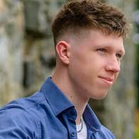 A gently smiling young man with blurry greenery in the background.
