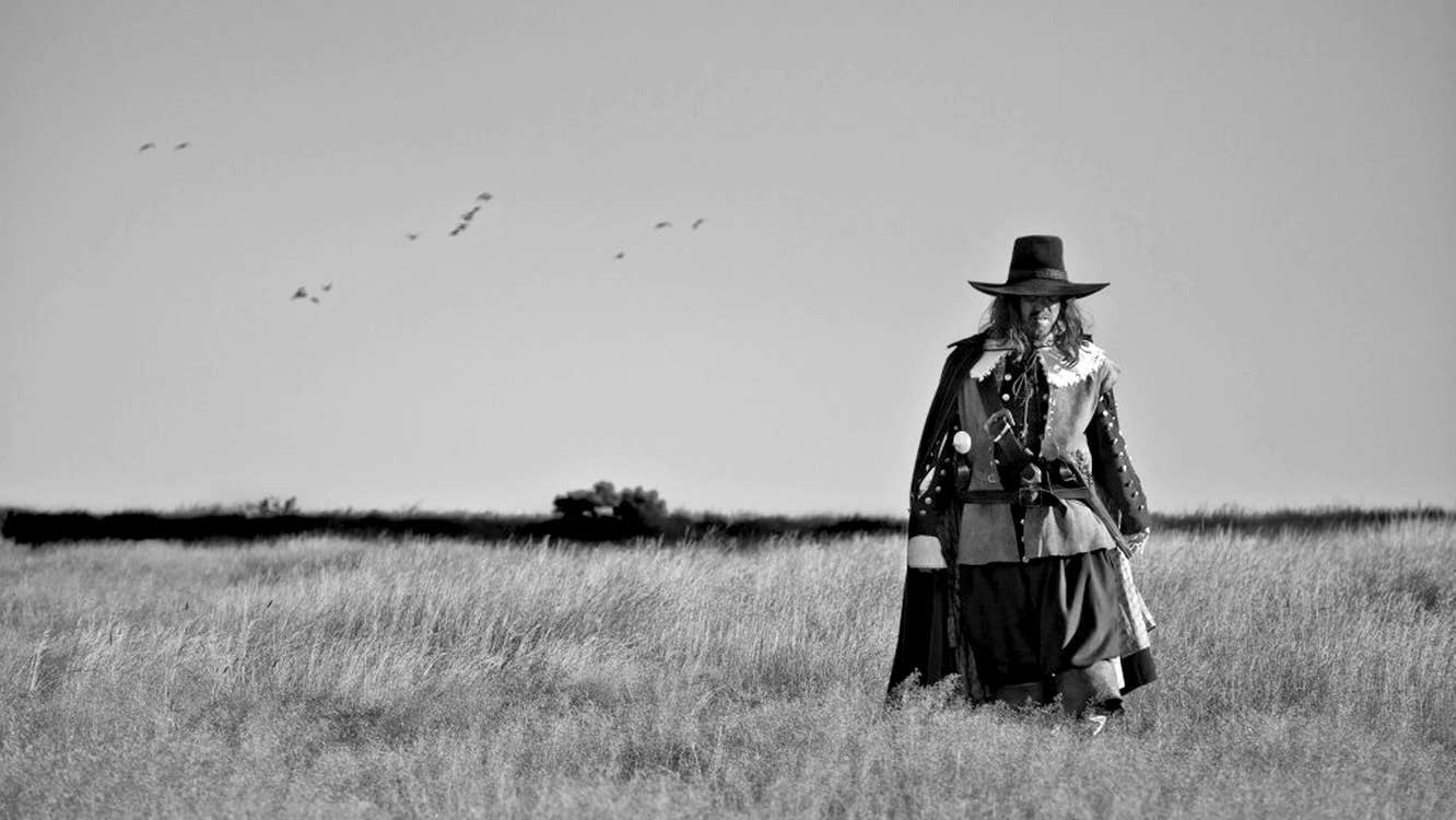 A man dressed in clothes of 17th Century England is walking across a field.