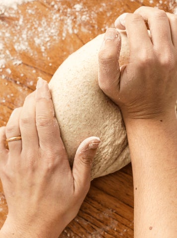 looking down on a pair of hands kneading a large piece of dough on a floured wooden surface.