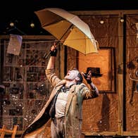 The Collector at An Táin, a man is holding an umbrella over his head on a stage surrounded by busy background stuff.