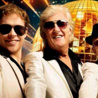 3 smiling men in white suits with dark shirts, all wearing sunglasses with golden, mosaic disco ball in background.