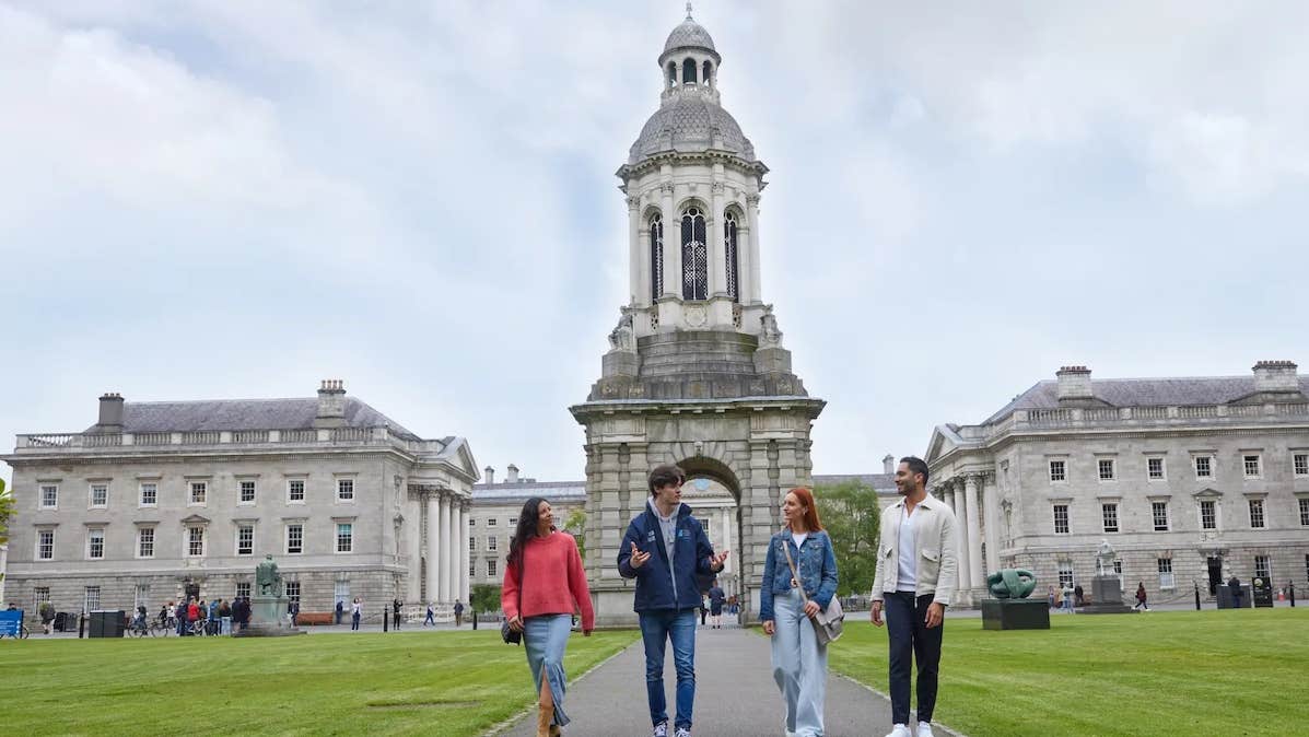 3 people walking on a path with a tour guide talking, impressive grand, stone buildings and tour in the background.