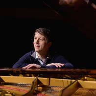 Looking across an open grand piano with a man sat at the keyboard end, looking away, resting his hands on top