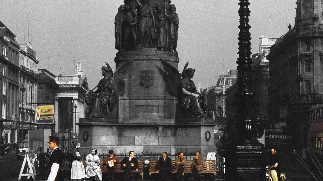 People scattered along 2 benches underneath plinth of large monument in a city in the past.