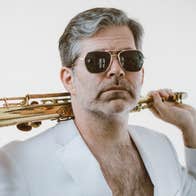 A man in sunglasses and a white open jacket showing a hairy chest is holding a gold coloured wind musical instrument over his shoulders.