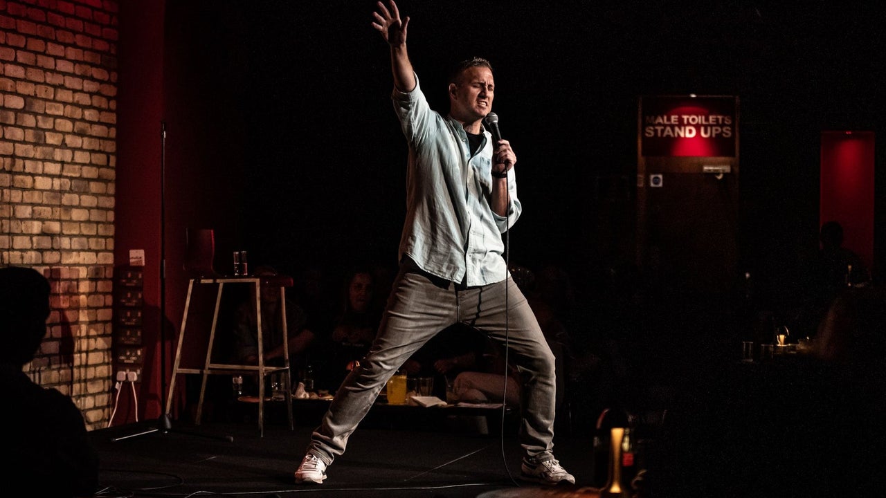 A comedian on stage holding a microphone with his right arm and leg extended