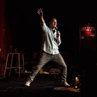 A comedian on stage holding a microphone with his right arm and leg extended