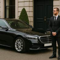 Person in a dark suit standing by a black car parked on a street