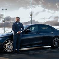 A driver stands beside a dark car in a parking area under a cloudy sky