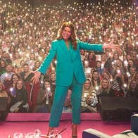 Doireann Garrihy - The Laughs Of Your Life Live, a smiling woman with arms outstretched on a stage with large audience in the background holding lights.
