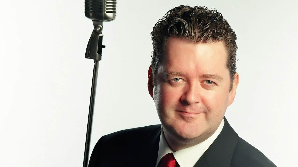 A man is smiling, in a dark jacket with red tie, beside is old fashioned style mic on a tall stand, against plain white background.