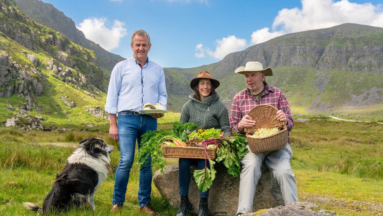 Three people posing with food in baskets and a sheepdog with a mountain in the background