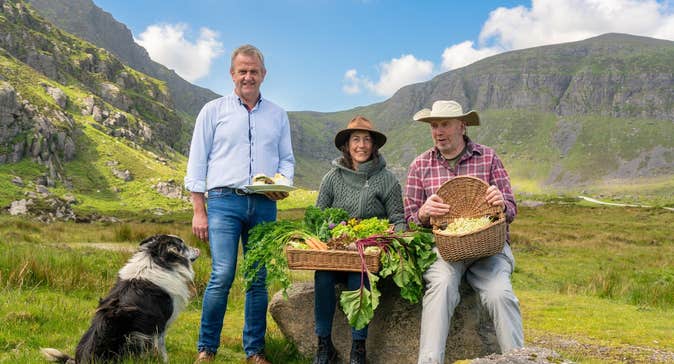 Three people posing with food in baskets and a sheepdog with a mountain in the background