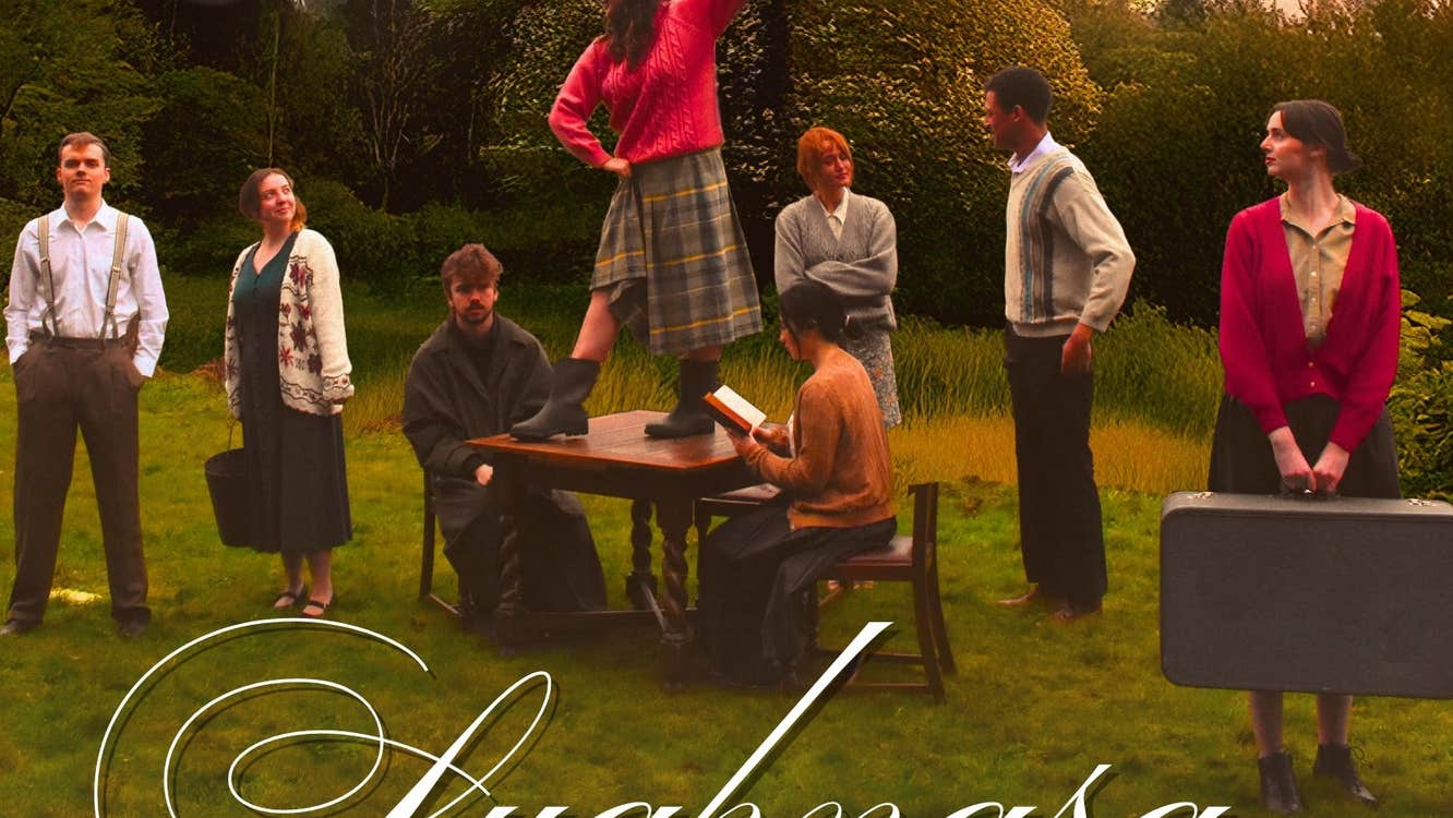 Dancing at Lughnasa - An Táin - A group of people standing outdoors on grass with 2 people seated at a table.