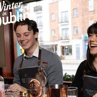 2 people are at a bar counter laughing with cocktail equipment in front of them.