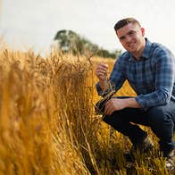 A smiling man crouched down beside golden wheat growing in a field.