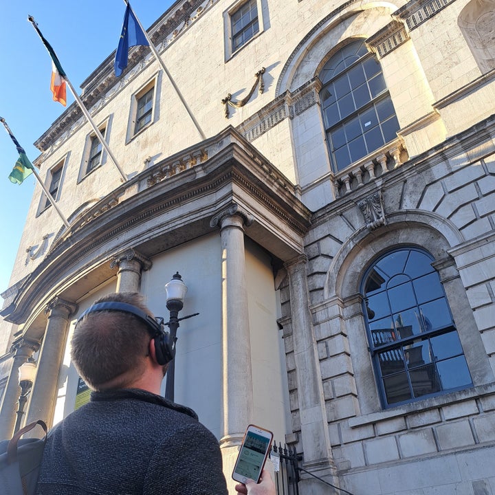 Person wearing headphones holding a phone looking up at a building with three flags flying