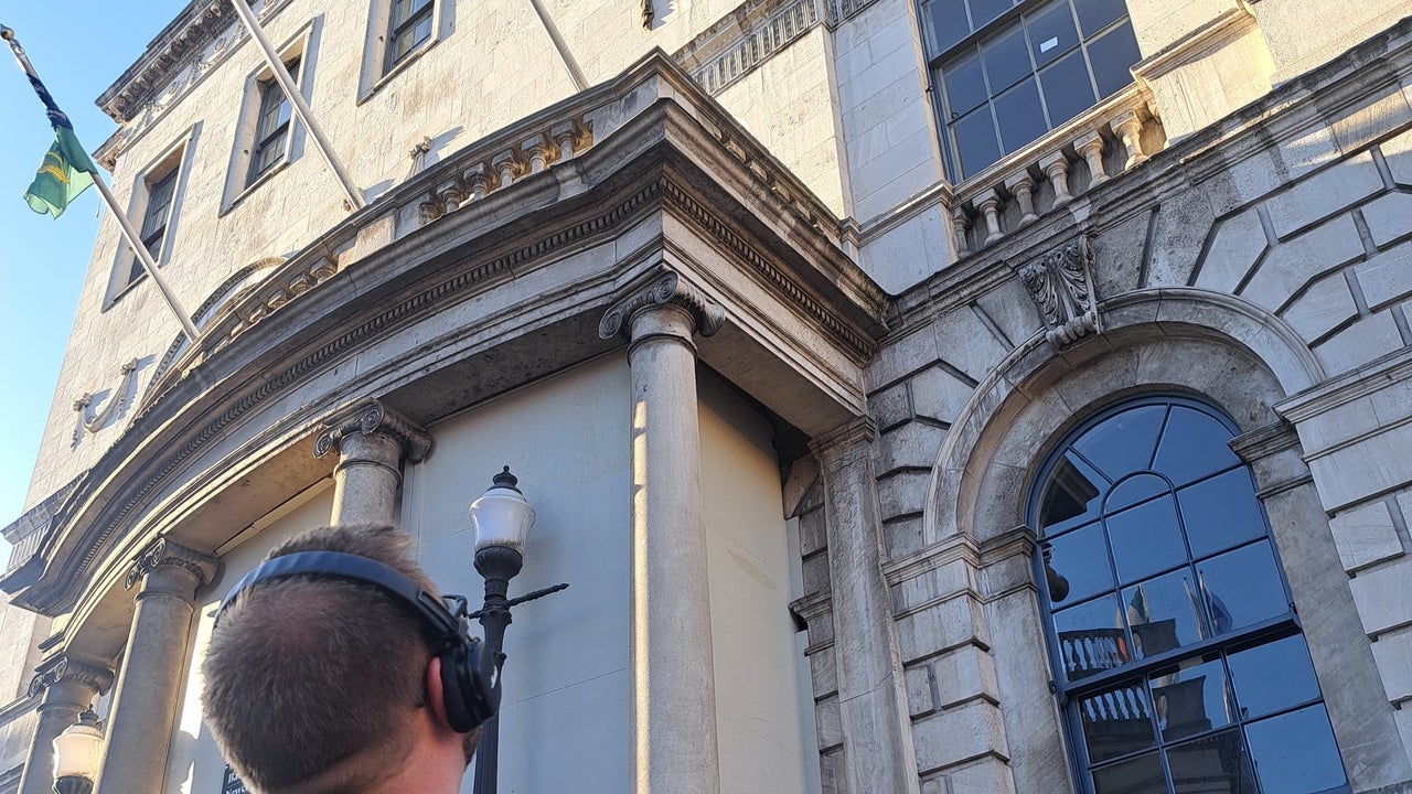 Person wearing headphones holding a phone looking up at a building with three flags flying