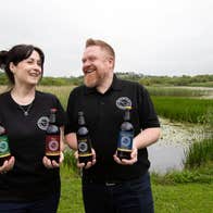 Two people showcasing a selection of beers against the backdrop of a lakeside wetland