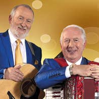 2 smiling older men resting their hands on musical instruments around their necks on straps