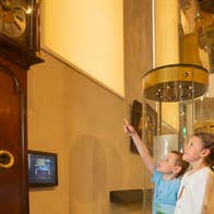 Two children looking up at a grandfather clock with one child is pointing up at it