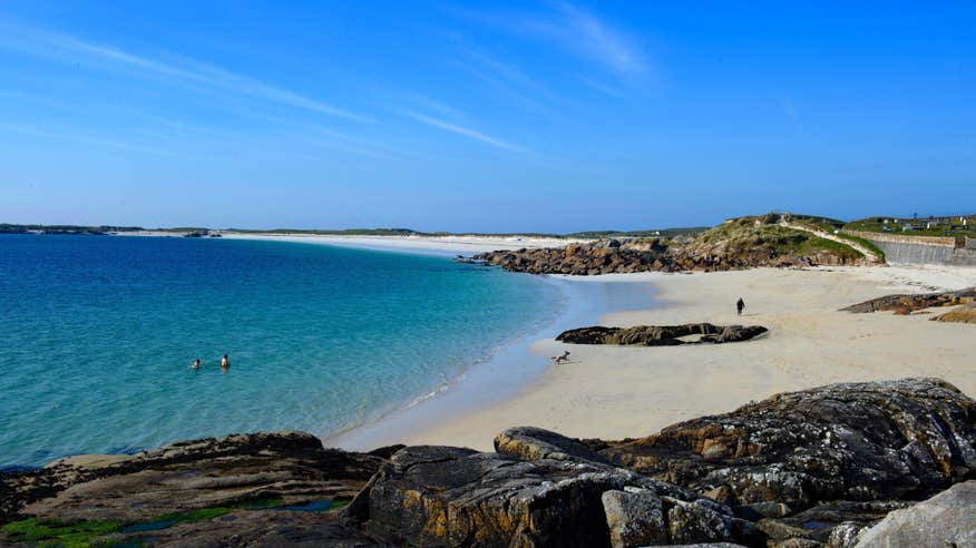 Two people bathing in clear blue water while a dog walks on the shore at Gurteen Beach in Galway