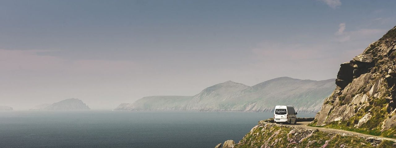 A minibus drives around a bend to the right by a cliff edge which opens out to the sea on the left