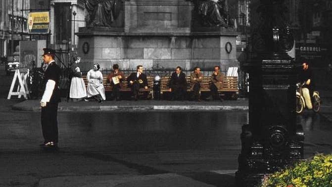 People scattered along 2 benches underneath plinth of large monument in a city in the past.